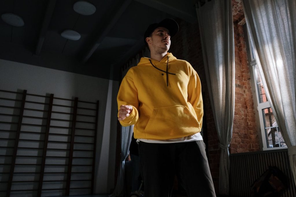 Man in yellow hoodie dancing indoors with dramatic lighting and shadows.